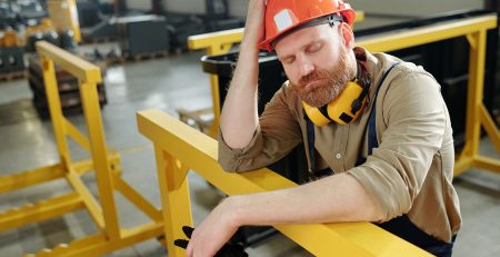 Tired male worker in hard hat leaning on railing on a factory floor.